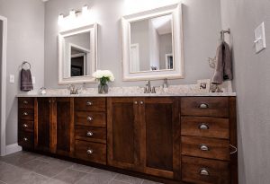 Modern bathroom with double sinks, large wooden vanity, granite countertop, two framed mirrors, wall-mounted lights, gray walls, and decorative items like flowers and towels.