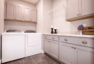 A modern laundry room with a washing machine and dryer, gray cabinets, a marble countertop, three decorative candles, a small potted plant, and a wire shelf above the machines. The floor is tiled in dark gray.