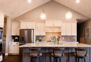 A modern kitchen with white cabinets, stainless steel appliances, a large island with four wooden chairs, pendant lights, and decorative items including flowers and plants on the countertop.