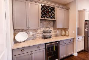 A modern kitchen with gray cabinets, a marble countertop, a built-in wine rack, a small drink fridge, a decorative plate, two glass vases, and a metal rack, against a stone tile backsplash.