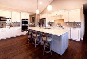 Spacious modern kitchen with white cabinets, stainless steel appliances, light granite island with three wooden stools, pendant lights, tile backsplash, and dark wood flooring. Flowers in a vase sit on the island.