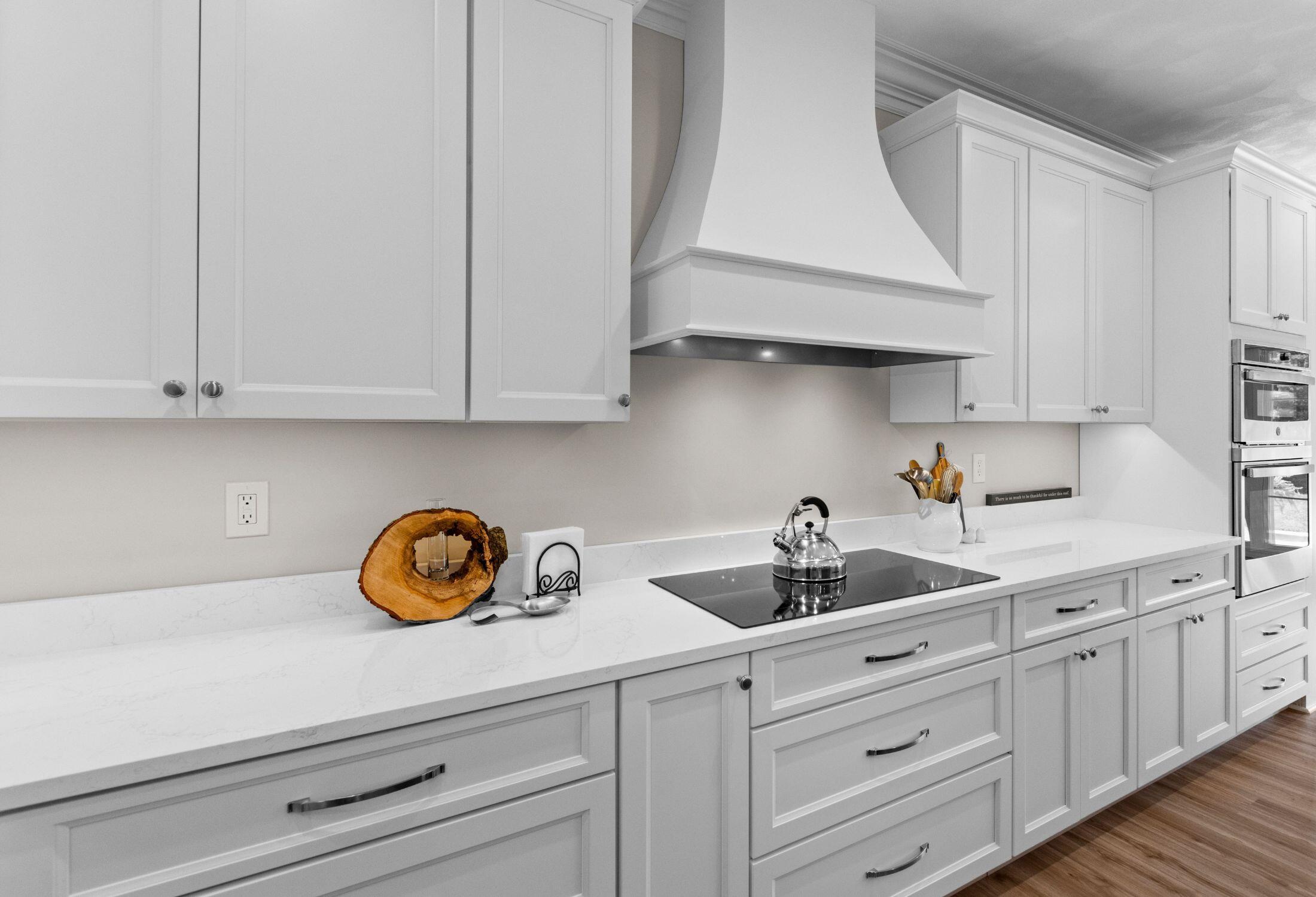 Modern kitchen with white cabinets and drawers, a marble countertop, electric stovetop with a kettle, built-in ovens, a large white vent hood, and decorative items including a wood slice and utensils in a jar.