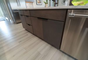 Close-up of modern kitchen cabinets with a dark wood finish, light-colored floor, and stainless steel dishwasher. Marble countertop, refrigerator, artwork, and a plant are visible in the background.