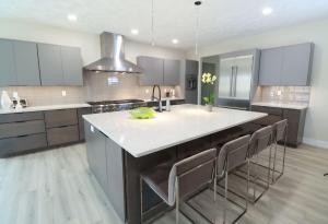 Modern kitchen with gray cabinets, stainless steel appliances, a large island with a white countertop, four bar stools, a sink, and a potted orchid. Light wood flooring and neutral backsplash complete the design.