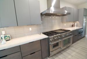 Modern kitchen with gray cabinets, stainless steel appliances, a large gas stove and oven, light tile backsplash, and white countertops. There is a range hood above the stove and a small framed picture on the counter.