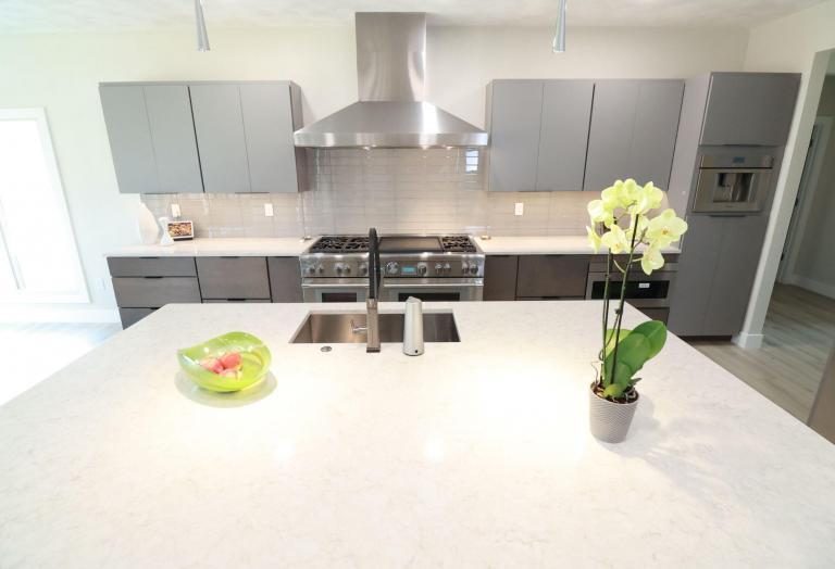 Modern kitchen with gray cabinets, stainless steel appliances, and a large island featuring a double sink, a green bowl of fruit, and a potted yellow orchid. The backsplash is light gray tile.