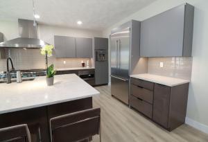 Modern kitchen with gray cabinets, stainless steel appliances, a large refrigerator, light wood flooring, a kitchen island with a potted orchid, bar stools, and under-cabinet lighting.