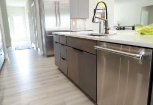 Modern kitchen with light wood flooring, stainless steel appliances, a large island with a sink and faucet, dark lower cabinets, and a bright, airy atmosphere. A green decorative bowl sits on the countertop.