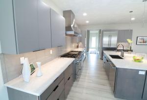 Modern kitchen with sleek gray cabinets, white countertops, stainless steel appliances, a large island with a sink, and bright lighting. Decorative items and a tablet are on the counters.